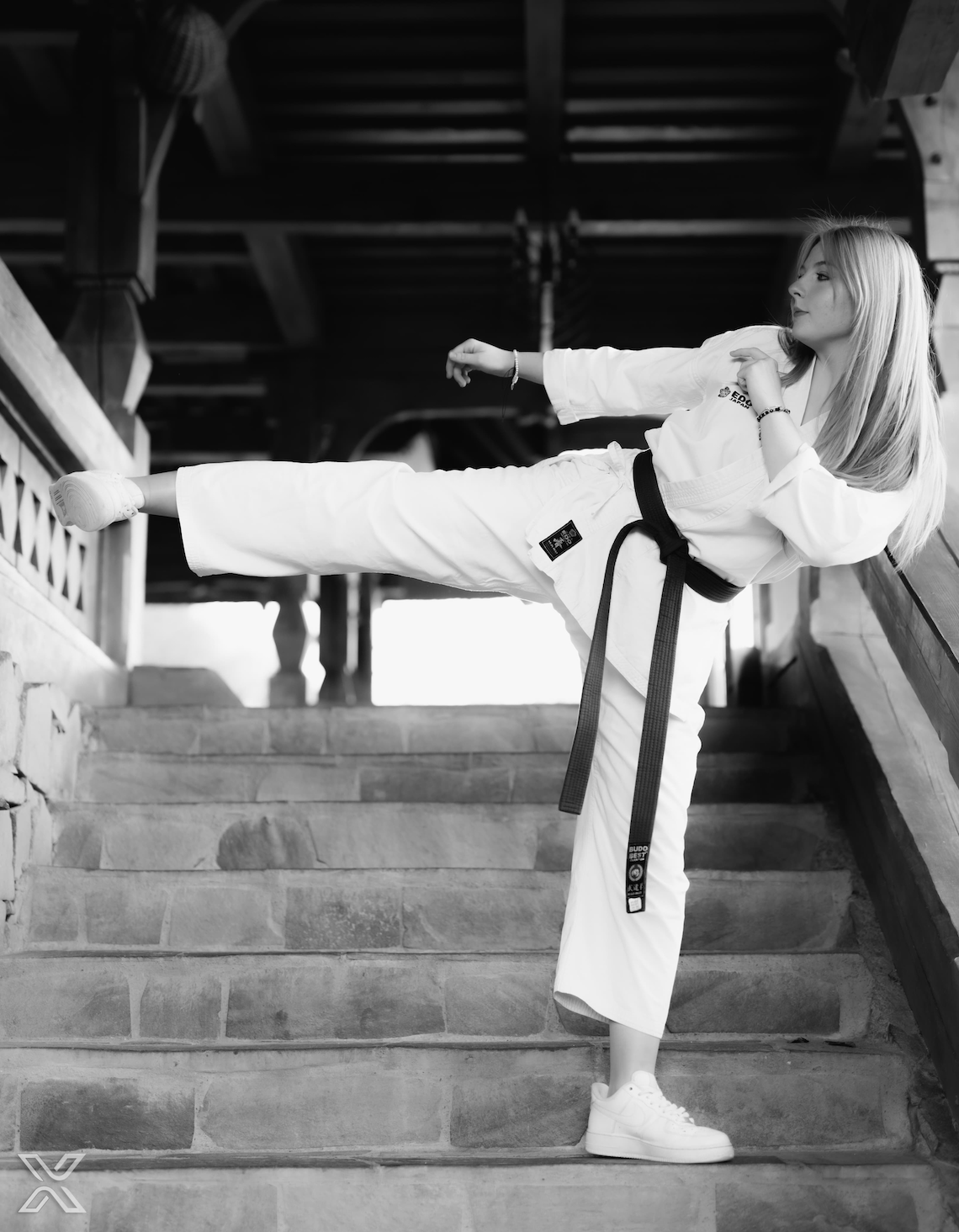 A young woman dressed in martial arts gear (kimono) with a black belt stands on a stone staircase in a traditional wooden building and performs a side kick with her right leg. The image is in black and white, highlighting her concentrated expression and strong posture.